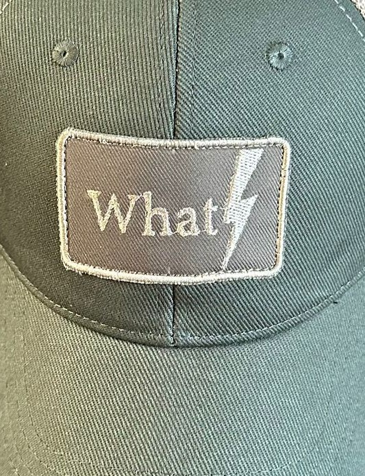 Detalle del parche What con rayo sobre fondo beis en la gorra Metro Cero de WhatStyle.